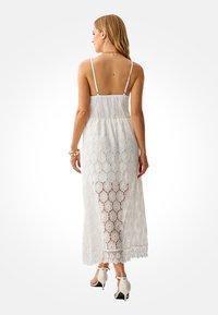 White lace maxi dress with adjustable thin straps, featuring a smocked bodice and floral pattern, finishing at the ankle with a scalloped hem.