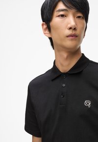 Čierna polo košeľa vyrobená z bavlny, s golierom, trojím gombíkovým zapínaním a malým bielym logom na ľavom prsníku. Hladká textúra.