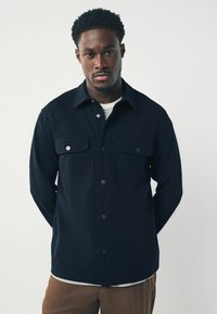 Camisa de botones azul marino hecha de tela suave, con dos bolsillos en el pecho con cierres de solapa y un cuello clásico. Debajo, se puede ver parcialmente una camisa blanca y azul a rayas.