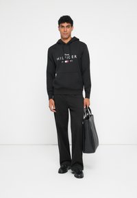 Fleece preto com logotipo branco e detalhes em vermelho e azul, combinado com calças pretas e sapatos pretos. Uma mala tote preta é segurada em uma das mãos.