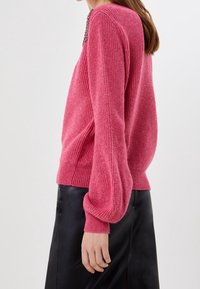 Žena v růžovém pleteném svetru s balónovými rukávy a černou koženou sukni, zachycena z boku na jednobarevném pozadí.