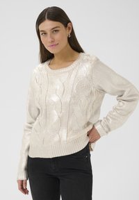 Maglione in maglia a treccia metallica con maniche lunghe, scollatura rotonda e orlo a coste. Indossato sopra jeans scuri, evidenziando il design strutturato.