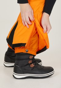 Svarta vattentäta stövlar med bruna accent-snören, parat med klara orange vattentäta byxor som har dragkedjor och elastiska muddar.