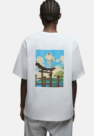 Personne aux cheveux tressés portant un T-shirt blanc montrant un lutteur de sumo faisant du skateboard au-dessus d'un portail torii japonais avec un ciel et des arbres en arrière-plan.