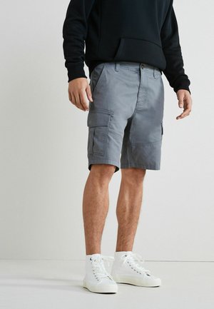 Pantalones cortos grises de cargo con múltiples bolsillos y un corte holgado, combinados con zapatillas altas blancas. El material parece duradero y ligero.