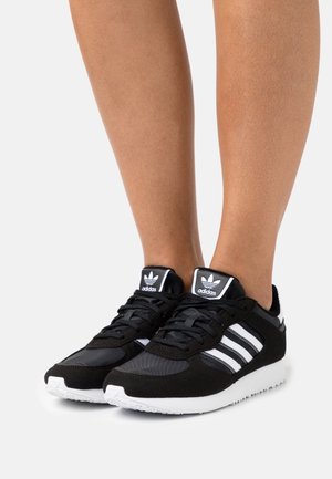 Zapatillas deportivas negras con rayas y detalles blancos, con un exterior de malla y gamuza, cordones gruesos negros y una suela de goma blanca.