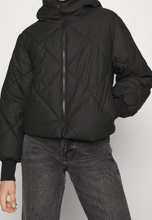 Kvinde iført en sort quiltet dunjakke med frontlynlås og høj krave, kombineret med falmede sorte jeans, stående med den ene hånd på hoften.