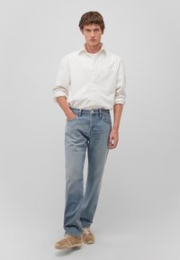 White long-sleeve shirt, blue denim jeans, and beige sneakers. Shirt has a single chest pocket; jeans are straight-leg with a faded finish.