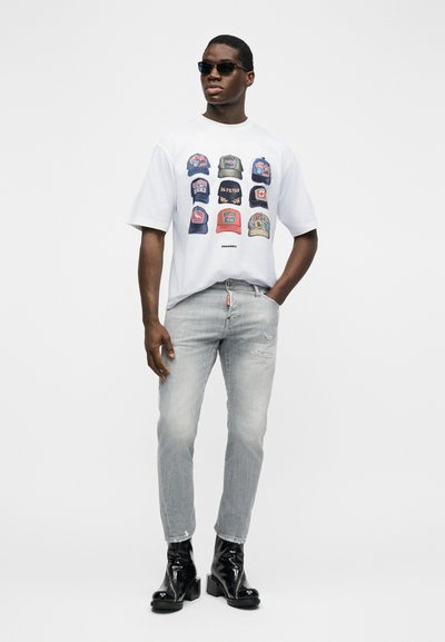 Άνδρας που φοράει γυαλιά ηλίου, λευκό γραφικό μπλουζάκι με εννέα πολύχρωμα σχέδια καπέλων, ανοιχτό γκρι τζιν και μαύρες γυαλιστερές μπότες μπροστά σε απλό φόντο.