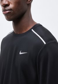 Άνδρας με μαύρο φούτερ Nike με λευκές ρίγες στους ώμους και λευκό λογότυπο Nike στο στήθος, σε απλό ανοιχτό φόντο.