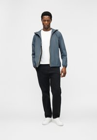 Νεαρός άνδρας με μπλε μπουφάν με κουκ ούλα πάνω από λευκό μπλουζάκι, μαύρο παντελόνι και άσπρα αθλητικά παπούτσια, στέκεται με τα χέρια στις τσέπες.