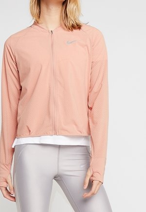 Person trägt eine hellrosa Sportjacke mit Reißverschluss und Daumenlöchern sowie hellgraue Leggings und steht vor einem einfarbigen Hintergrund.
