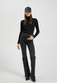 Schwarzer gerippter Rollkragenpullover kombiniert mit hochtaillierten schwarzen Schlagjeans; das Model trägt eine schwarze Mütze und spitze schwarze High Heels.