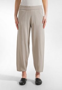 Henkilö seisoo päällään löysät beigestä väristä valmistetut housut, joissa on leveä ribbineulottu vyötärönauha, mustat slip-on-kengät ja valkoinen tausta.