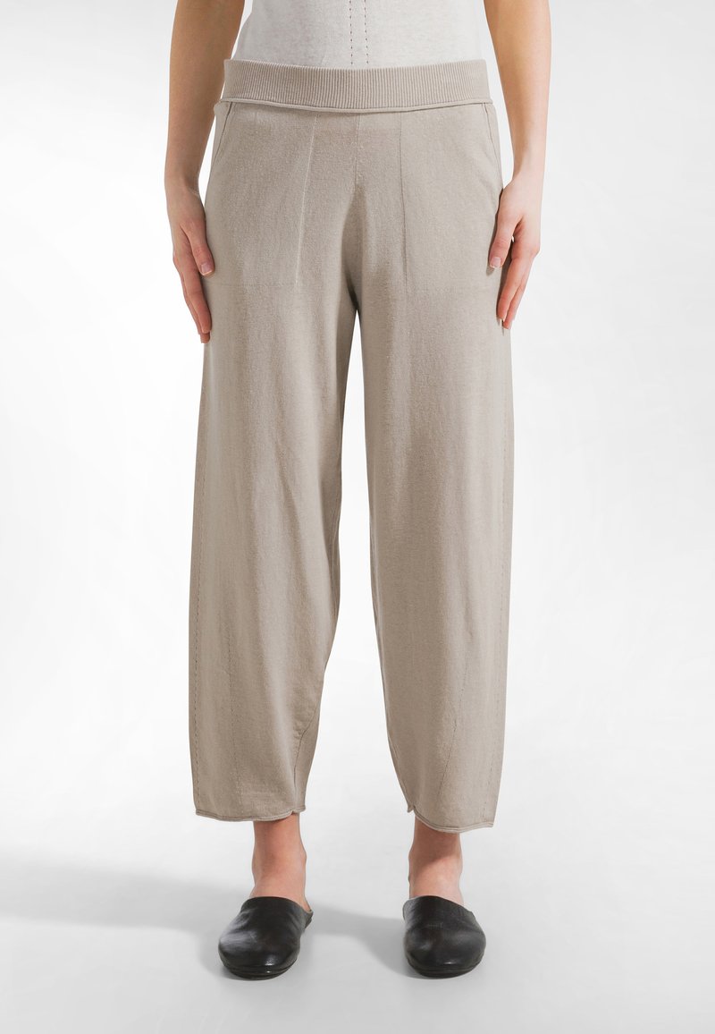 Henkilö seisoo päällään löysät beigestä väristä valmistetut housut, joissa on leveä ribbineulottu vyötärönauha, mustat slip-on-kengät ja valkoinen tausta.