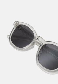 Klare Kunststoffsonnenbrille mit dunklen Gläsern, ausgestattet mit einem massiven Rahmen und runder Form. Auffällige Akzente sind ein dekoratives Element an den Scharnieren.