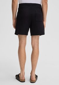 Zwarte shorts van textuurstof, met een elastische tailleband en een enkele achterzak. Draagt zwarte slidesandalen.