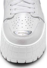 Baltās ādas Air Jordan sporta apaviem ir teksturēts augšdaļa, perforēta pirkstu kaste un bieza, rievota balta zole ar sudraba logo plāksni.
