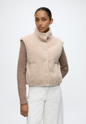 Donna che indossa un gilet sherpa beige senza maniche sopra un maglione marrone a maniche lunghe e pantaloni bianchi, in piedi davanti a uno sfondo chiaro e uniforme.