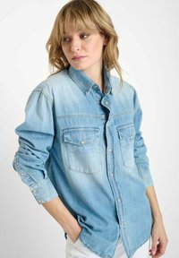 Lichtblauwe denimshirt met lange mouwen, knoopsluiting aan de voorkant, twee borstzakken en een zachte, vervaagde textuur. De klassieke kraag voegt detail toe.