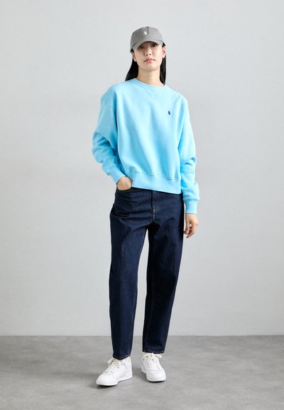 Felpa azzurra chiara con polsini a coste, jeans blu scuro e sneakers bianche. Cappellino grigio con dettagli del logo. Stile semplice e casual.