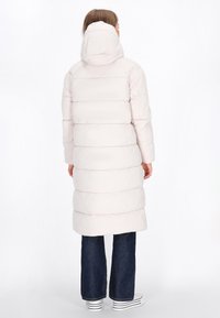 Lichtroze, lange gewatteerde jas met een capuchon, voorzien van horizontale quilted secties en zijzakken. Gecombineerd met donkere spijkerbroeken en sneakers.