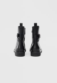 Bottes noires en cuir avec une finition lisse, dotées d'une sangle latérale et de détails en boucle, ainsi qu'une semelle en caoutchouc basse. Logo sur le talon.