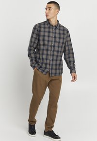 Langärmliges karierter Hemd in Marineblau, Braun und Weiß, mit Knöpfen und einem entspannten Schnitt, kombiniert mit braunen Hosen und schwarzen Sneakers.