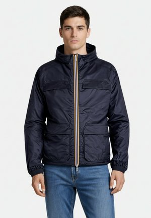 Mężczyzna noszący granatową kurtkę z kapturem z przednimi kieszeniami i suwakiem, w połączeniu z niebieskimi dżinsami, stoi na białym tle.