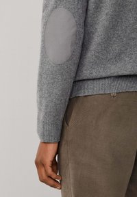 Maglione grigio con orlo e polsini a costine; presenta toppe per i gomiti grigie e lisce. Indossato sopra pantaloni in velluto a coste marroni, che mostrano una finitura testurizzata.