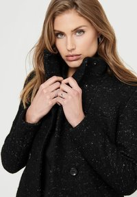 Manteau noir en mélange de laine avec une finition texturée, à double boutonnage et col montant. Présente de grands boutons et des détails de couture visibles.