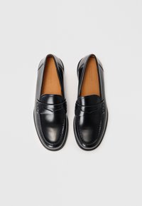 Mustad nahast loafers ümmarguse toe, dekoratiivse õmblemise ja väikese kõrgendusega aktsendiga rihmal. Interjöör on kaetud beežist voodriga.