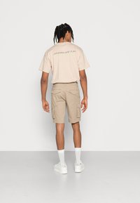 Beige �übergroßes T-Shirt mit grafischem Text auf der Rückseite, kombiniert mit khakifarbenen Cargo-Shorts mit seitlichen Taschen, getragen mit weißen Sneakers und Socken.