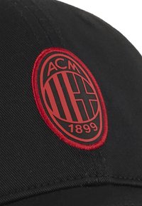 Svart cap med en rund, broderad röd logotyp med svarta accenter, som visar bokstäverna "ACM" och året "1899."