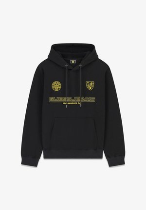 Sudadera con capucha negra con texto amarillo "GUESS JEANS Los Angeles, CA" y dos logotipos amarillos, uno circular y otro en forma de escudo, en el pecho, bolsillo delantero y cordones.