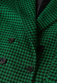 Blazer vert et noir à motif pied-de-poule avec boutons noirs, montrant le revers et les détails de la fermeture double boutonnage.
