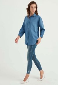 Camicia blu con bottoni, dalla vestibilità rilassata, abbinata a jeans blu strappati e scarpe con tacco bianco. Modella sta camminando.