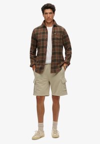 Flanellen shirt in bruine en groene ruit met een knoopkraag, witte T-shirt, beige cargoshorts en beige sneakers met zichtbare veters.