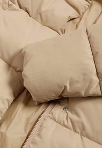 Beige PUFFERTJEN med quiltet design, blød tekstur og sølv lynlås. Har polstrede manchetter og trykknapdetaljer.