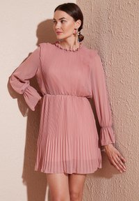 Roza plisirana haljina s dugim rukavima, volanastim izrezom i elastičnom maramom. Prozirna tkanina dodaje teksturu. Model pozira uz zid.