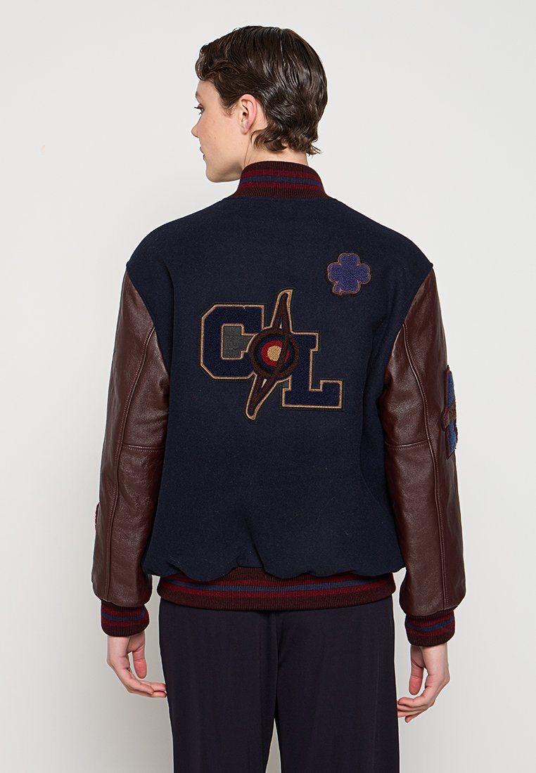 Giacca varsity in lana navy con maniche in pelle marrone, colletto a coste e orlo ondulato. Presenta loghi ricamati e toppe floreali.