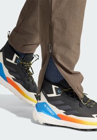 Μαύρα ορειβατικά παπούτσια με κίτρινες και μπλε λεπτομέρειες, σε συνδυασμό με καφέ παντελόνια με φερμουάρ και υφή. Οι κάλτσες είναι σκούρο μπλε.