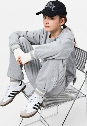 Grijze sweatshirt en broek met witte en grijze gestreepte ribbels, gecombineerd met witte sneakers met zwarte accenten. Een zwarte pet maakt de look af.