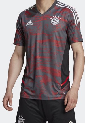 Mężczyzna ubrany w szarą i czerwoną wzorzystą koszulkę piłkarską Adidas FC Bayern Monachium z białymi paskami na ramionach i pasujące czarne spodenki.