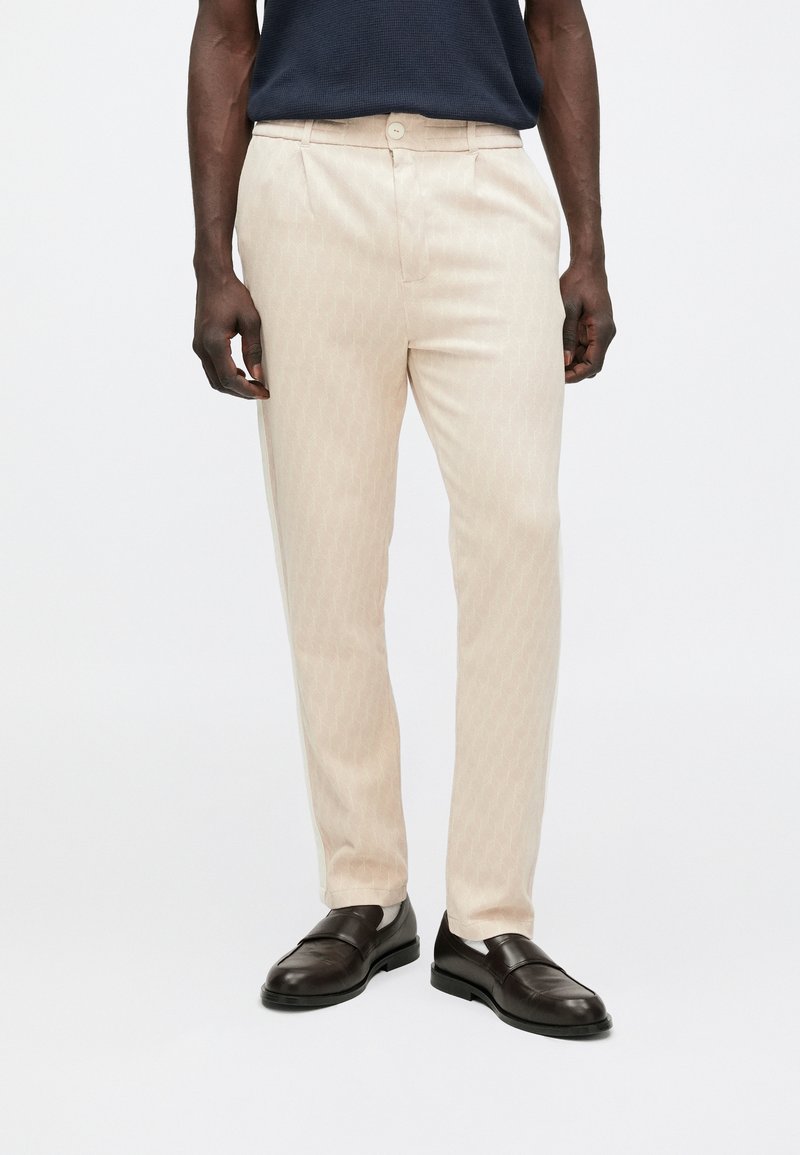 Beige mønstrede bukser med rett passform, med sidelommer, beltestropper og knappelukking, kombinert med mørke sko.