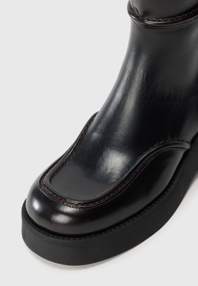 Botte en cuir noire avec une finition lisse, bout arrondi, couture contrastante en brun et semelle en caoutchouc épaisse pour une meilleure adhérence.
