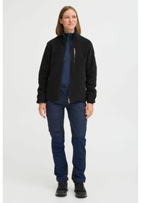 Veste en polaire noire avec poche poitrine zippée, associée à un pantalon cargo bleu marine doté de poches latérales et d'une coupe fuselée. Des chaussures noires complètent la tenue.