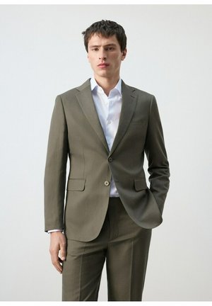 Uomo con indosso un abito verde oliva e una camicia bianca, in piedi con una mano in tasca su sfondo uniforme.