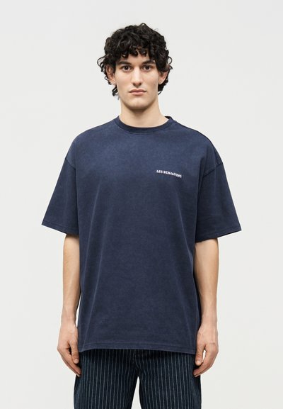 Jeune homme aux cheveux bouclés et foncés, portant un t-shirt ample bleu foncé avec un petit texte blanc sur la poitrine et un pantalon à fines rayures, debout devant un fond blanc.