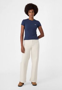 Marineblauw T-shirt met korte mouwen en een klein wit logo op de borst, gecombineerd met crèmekleurige wijde broekspijpen en bruine schoenen.